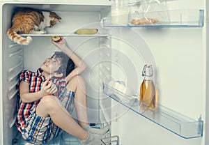 A boy in a shirt and shorts playing with red cat inside a fridge