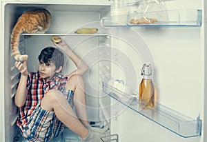 A boy in a shirt and shorts with red cat inside a fridge. A boy is going to take a cat tail