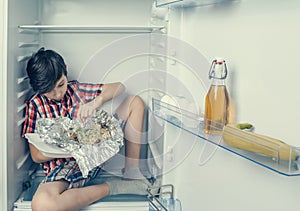 A boy in a shirt and shorts unwrapping a food package sitting in a refrigerator. Close-up