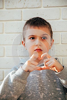 The boy shows a heart with his fingers. Portrait of a child against a white brick wall