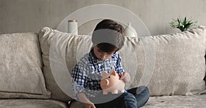 Boy sit on sofa counts coins throwing them into piggybank