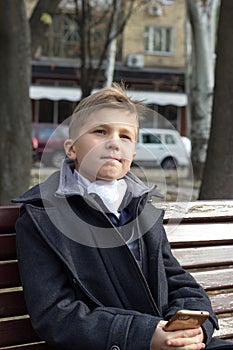 A boy sits on a bench in a park in business clothes with an appraising look and a slight smile. Thoughtfulness on his photo