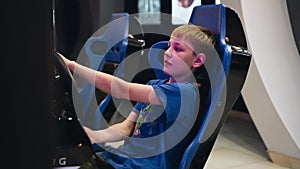 The boy sits in a chair racing car playing a video game. The view from the side. The boy`s face shows emotion.