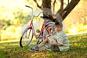 The boy sits on the grass in the garden and thinks about the book read.  A red bicycle stands by a tree, a book is lying on the