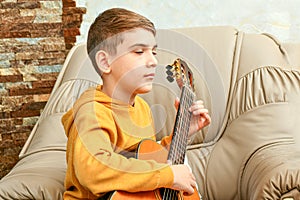 The boy sits at home on a leather armchair and plays a six-strin
