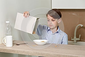 The boy sits in the kitchen at the table and prepares a breakfast of milk