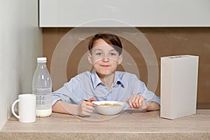 The boy sits in the kitchen at the table and prepares a breakfast of milk