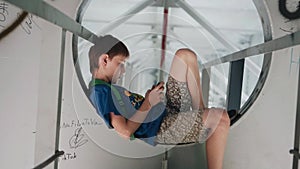 The boy sits on metal structures under the bridge. Walk around town. Urban environment. Modern society.