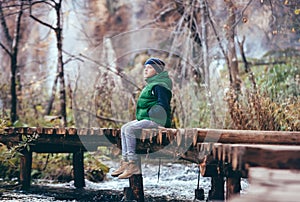 Boy sits omn wooden bridge over river in mountain autumn forest