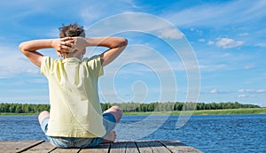 The boy sits on the pier, stretches and breathes air, against the lake, on the right there is a place for the inscription