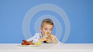 The boy sits at the table eats fast food and rejoices on a blue background. The concept of healthy and unhealthy food