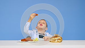 The boy sits at the table eats fast food and rejoices on a blue background. The concept of healthy and unhealthy food