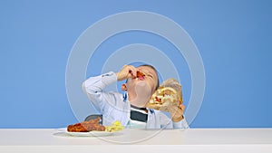 The boy sits at the table eats fast food and rejoices on a blue background. The concept of healthy and unhealthy food