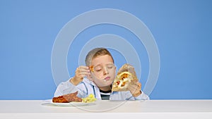 The boy sits at the table eats fast food and rejoices on a blue background. The concept of healthy and unhealthy food