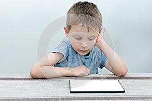 The boy sits at the table and looks at the tablet with interest, concentration and seriousness. Isolated screen for