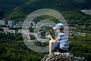 The boy sits on the top of the mountain and enjoys a panoramic view of the verdant valley and the city of Zhigulevsk.