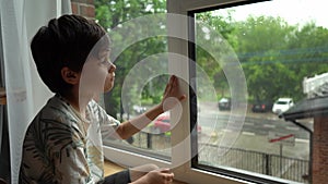 A boy sits at a wet window during the rain and looks at the street