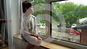 A boy sits at a wet window during the rain and looks at the street