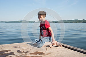 The boy sits on a wooden pier and looks at the water of the lake on a sunny day