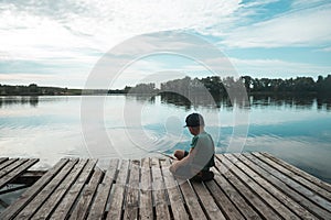 Boy sitting alone on pier while fishing from dock on lake. Kid on beautiful morning in nature