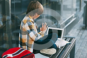 The boy is sitting on a bench, next to a laptop and a backpack. The boy communicates with friends using the computer, waving to
