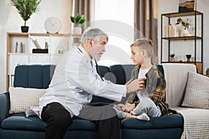 Boy sitting on the couch while caring experienced nice mature male doctor listening his heartbeat