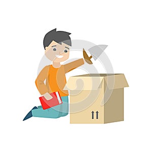 A boy is sitting on the floor and packing his toy sail-boat and a red book in a cardboard box