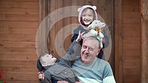 Boy sitting on grandfather knees, girl standing behind and hug. Brother and sister playing with toy sheep, laughing. 4K