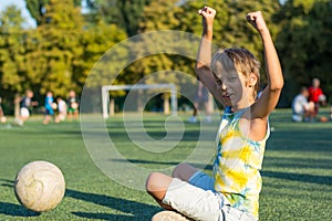 A boy is sitting on the grass at the edge of a stadium, watching football and rejoices at the soccer ball