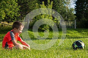 The boy is sitting on the grass and looks at the soccer ball