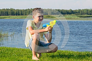 Boy sitting on green grass and playing with a water pistol is configured on the target on the background of beautiful scenery