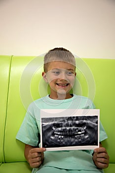 A boy is sitting on a green sofa in a dental clinic with a picture of his teeth in his hands. Boy with an 3d x-ray