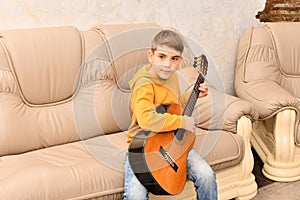 The boy is sitting on a leather sofa and playing the guitar