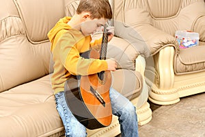 The boy is sitting on a leather sofa and playing the guitar