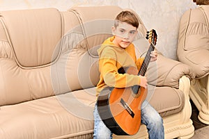 The boy is sitting on a leather sofa and playing the guitar