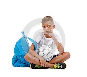 A boy sitting in the lotus position. A sportive kid with bright satchel and soccer ball isolated on a white background.