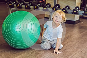 The boy is sitting near the fitball in the gym