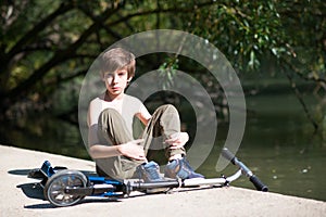 Boy sitting and resting on the riverbank with his scooter, summer nature outdoor.