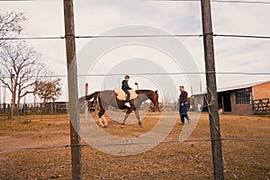 A boy, sitting on a saddle and riding a brown-furred horse at a stable with a stable boy, outdoors