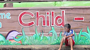 Boy sitting on sidewalk pavement of Mural painted concrete school wall waiting for his playmates.