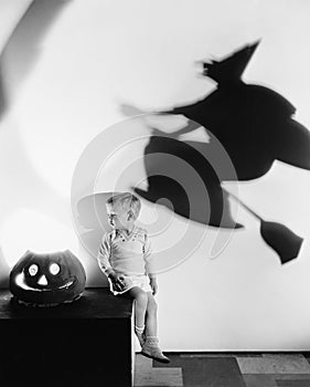 Boy sitting on a table with a pumpkin and a shadow of a witch on a wall