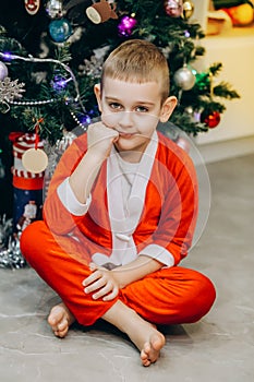 The boy is sitting under the Christmas tree in a Santa costume without a hat, looking at the camera with his chin propped on his