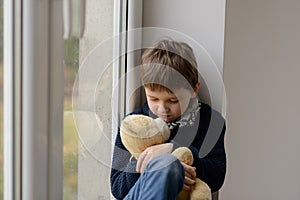 Boy sitting on the windowsill and hugs his favourite teddy bear