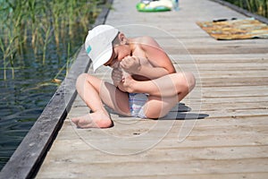 A boy sitting on a wooden floor, teeth pulls a thorn from his foot.