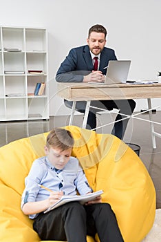 Boy sitting on yellow pillow and drawing while his father businessman