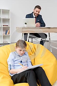 Boy sitting on yellow pillow and drawing while his father businessman