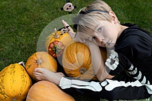 boy in a skeleton costume sits near many orange pumpkins, with a spider hanging from his mouth