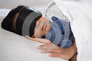 Boy sleeping on bed with teddy bear white pillow and sheets wearing sleep mask