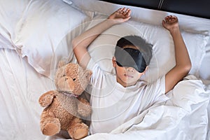 Boy sleeping on bed with teddy bear white pillow and sheets wearing sleep mask