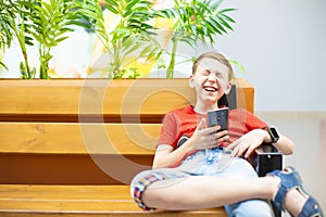A boy with a smartphone and smart clock sits on a bench and looks into the phone and laughs hard.